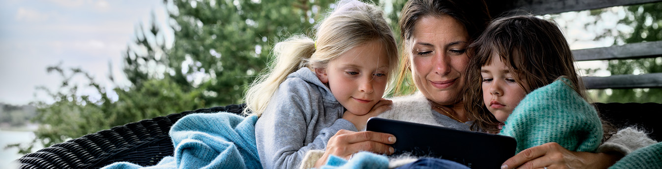 En kvinne sitter på terrassen med to små barn og ser på et nettbrett